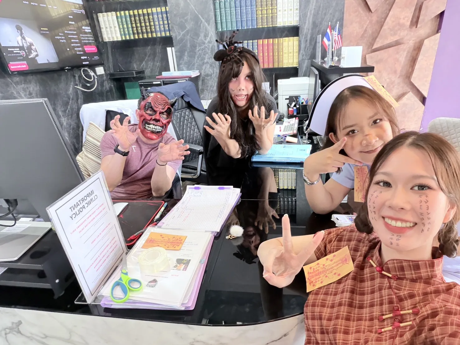 Four Maximum Clinic Bangkok team members in Halloween costumes making playful poses at the front desk: one in a red devil mask, one in spooky makeup with a spider headband, one in a nurse costume, and one in a red plaid qipao with Chinese jiangshi-style face markings and a paper talisman. Bookshelves, Thai and US flags and a clinic policy sign are in the background.