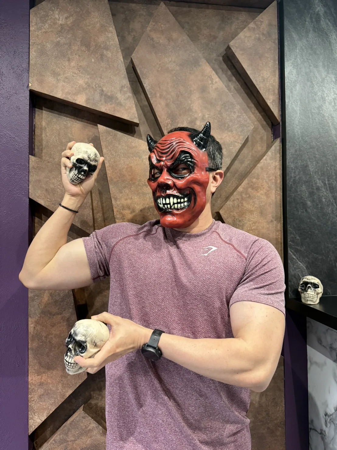 Dr. Krit in a horned red devil mask and a maroon athletic T-shirt, flexing one arm while holding a toy skull in each hand, in front of a bronze geometric wall at the Maximum Clinic Bangkok office