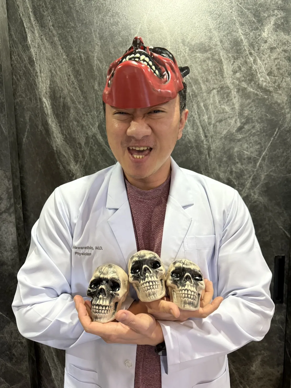 Dr. Krit in a white physician's coat with the red devil mask pushed up on his head, grinning widely and holding three toy skulls against a dark marble wall